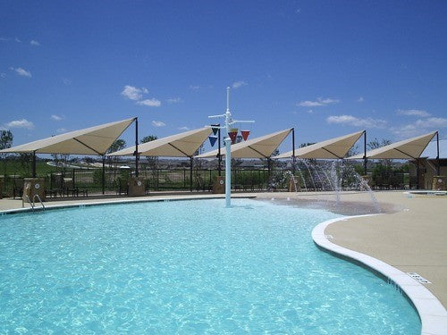 Wave-shaped beige fabric shade structure with four posts along a poolside under clear blue sky