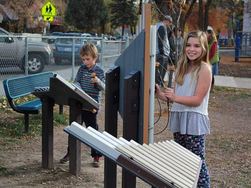 Two children playing on outdoor musical park instruments including a xylophone and metal chimes in a playground setting