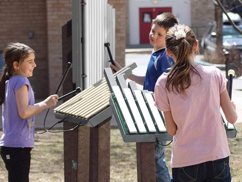 Three children playing on outdoor metal bar musical instruments with mallets, engaging in group music activity at a playground.