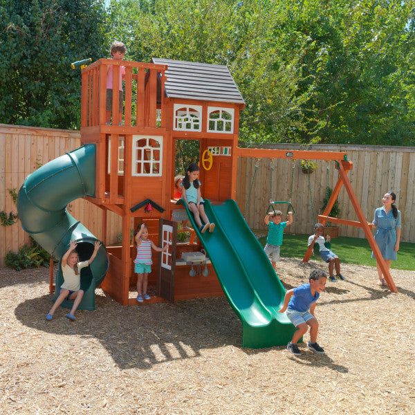 Wooden playset with double green slide, spiral tube slide, swings, climbing ladder, and children playing outdoors