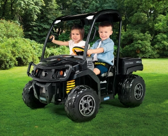 Black John Deere Gator XUV ride-on vehicle with roof, large wheels, and two children seated on grass outdoors