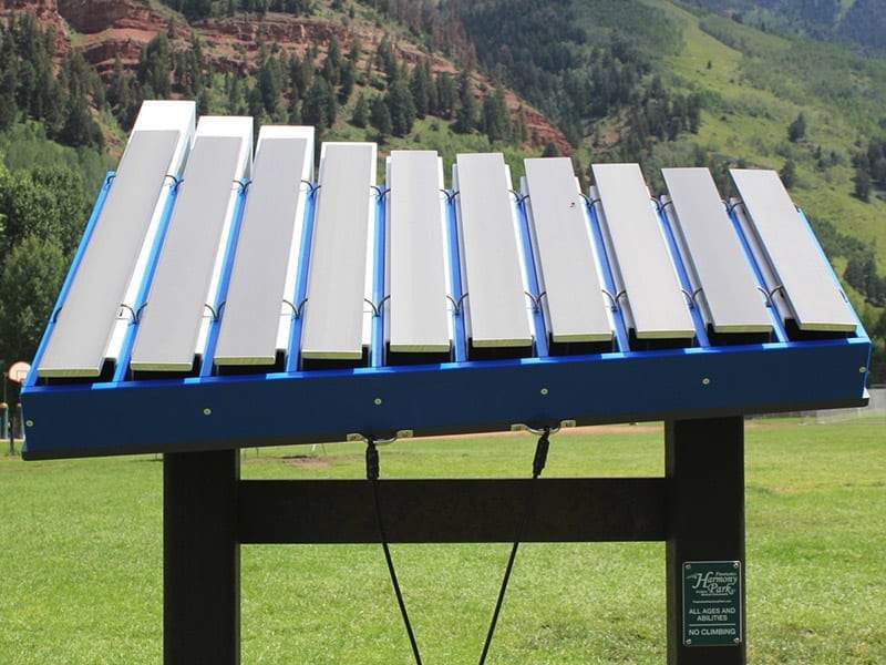 Outdoor blue musical instrument with eleven aluminum bars on a stand in a grassy park with forested hills.