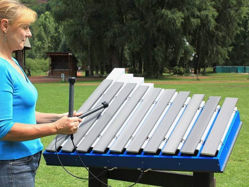 Woman playing large blue outdoor metallophone with silver aluminum bars in park setting