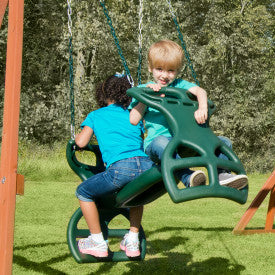 Green dual rider swing with two children seated, featuring handles and footrests, in an outdoor grassy play area