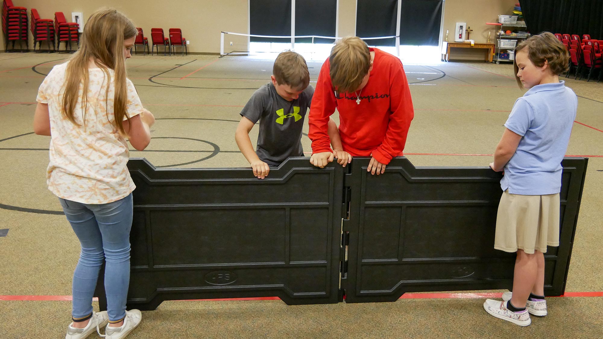Four children assembling black foldable panels for a portable indoor GaGa ball pit in a gym setting