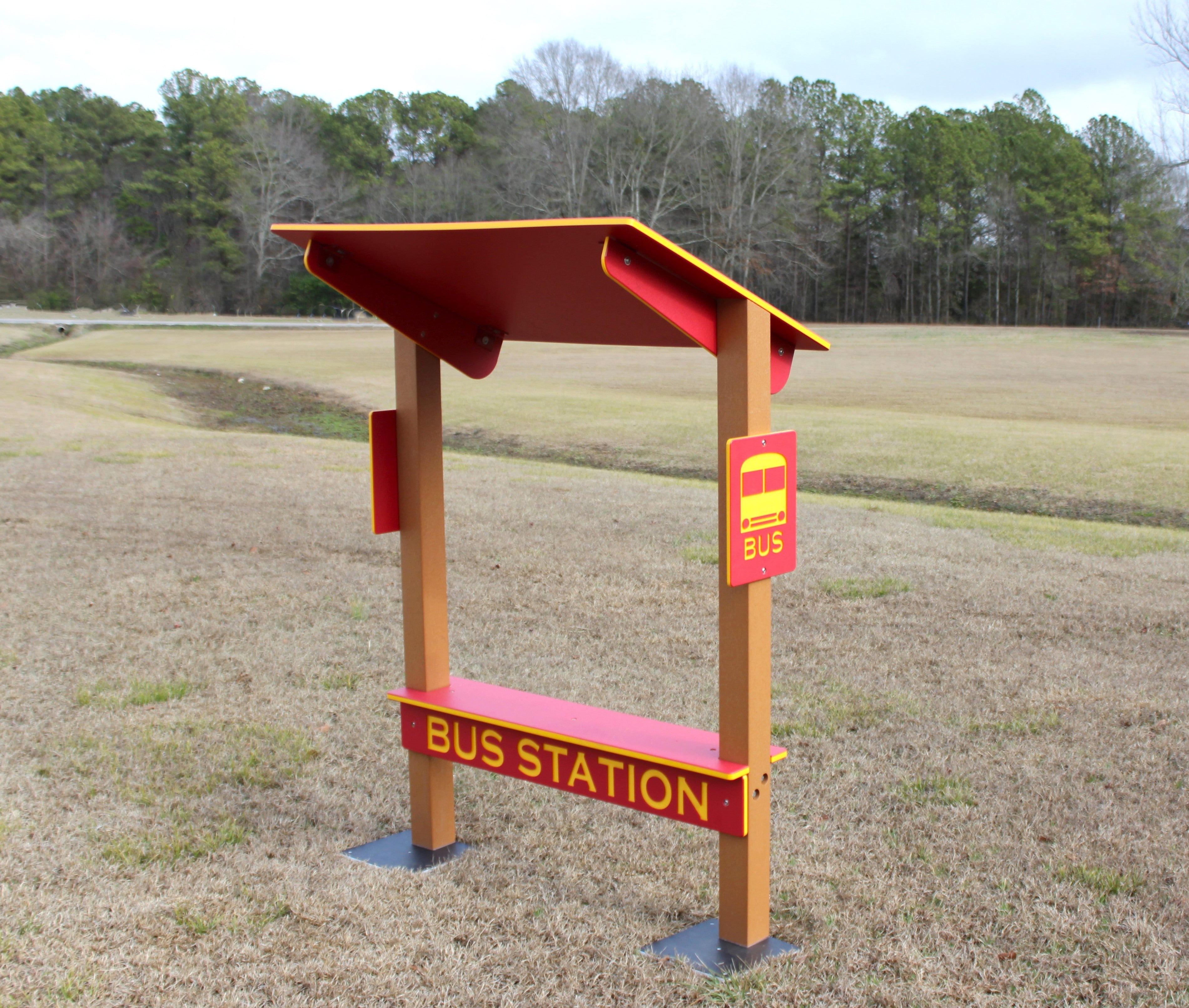 Red and yellow outdoor bus station bench with shaded roof, bus icon sign, and sturdy wooden posts in grassy play area