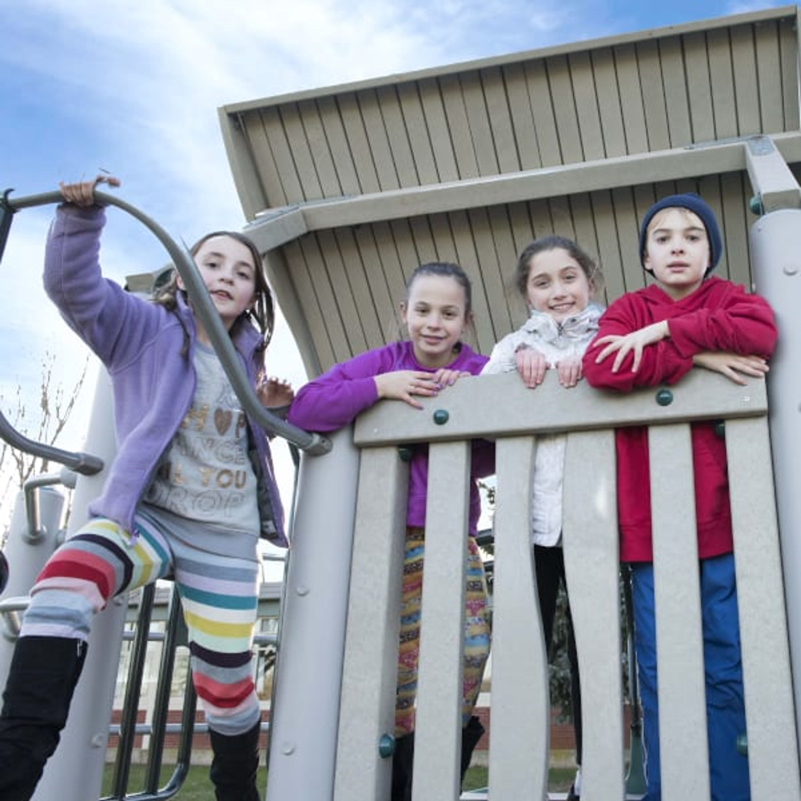 Four children in colorful jackets leaning on a beige railing under a roofed platform of an ADA-accessible playground structure