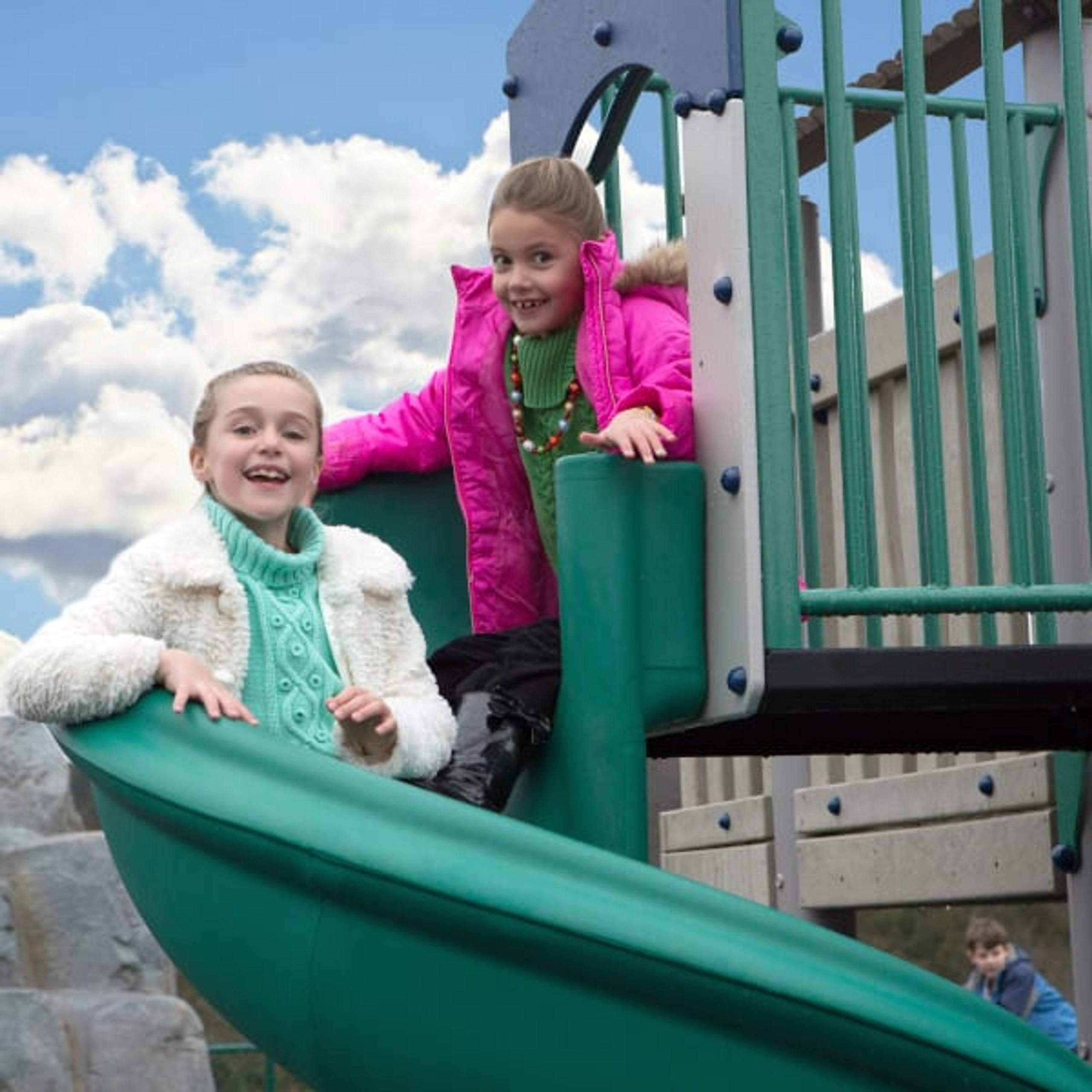 Two children playing on a green spiral slide attached to a wooden playground structure with climbing features
