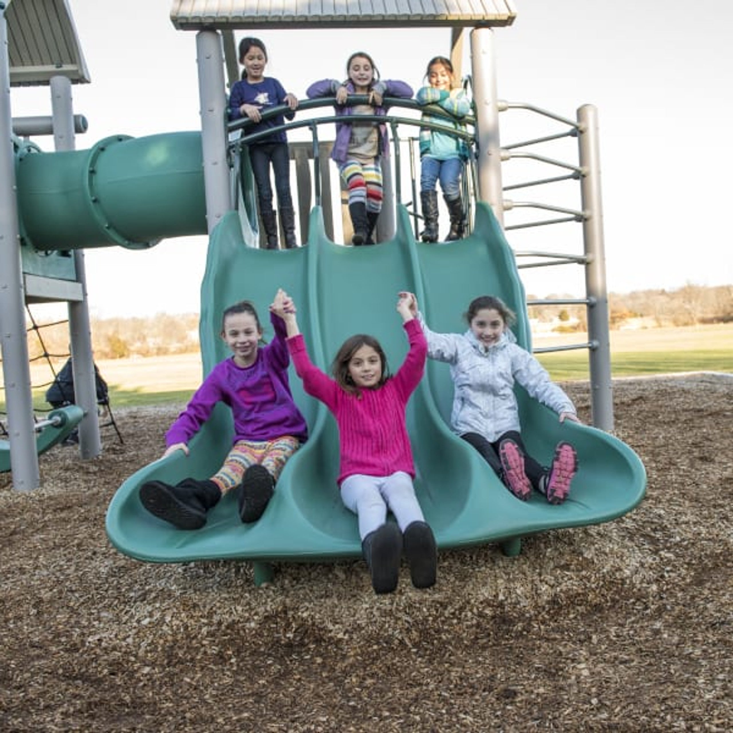 Triple wave slide on a large playground structure with children sliding and standing, featuring climbing elements and accessible design.